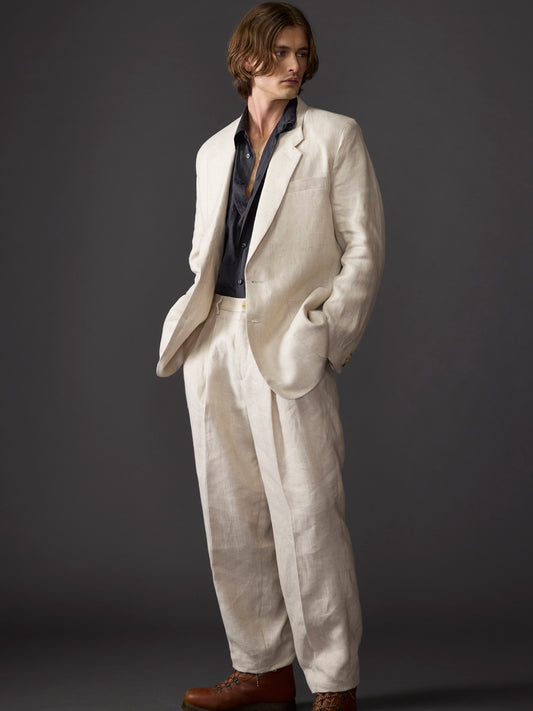 Full length image of a male model with chin-length hair wearing a relaxed fit beige linen suit featuring 1990s-inspired design details, set against a dark background.