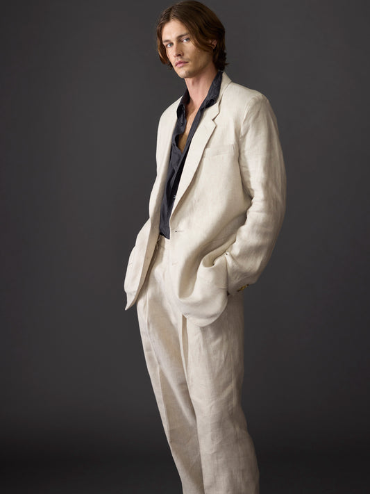 Male model with chin-length hair wearing a relaxed fit beige linen suit, showcasing the 1990s-inspired design, against a dark background.