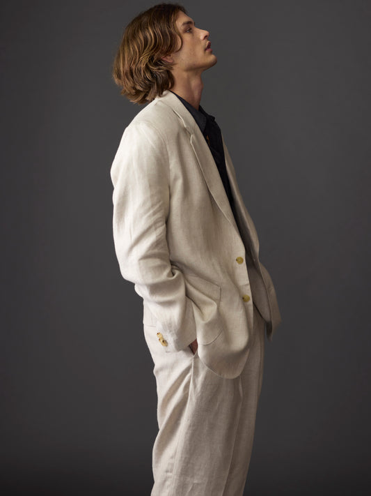 Cropped side view of a male model with chin-length hair wearing a relaxed fit beige linen suit featuring 1990s-inspired design details, set against a dark background.