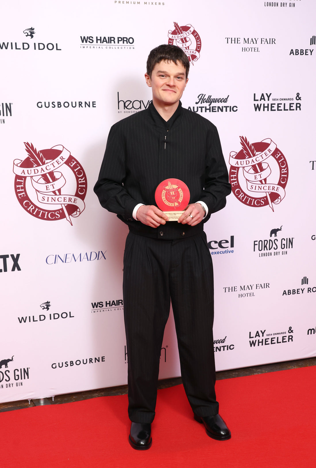 Robert Aramayo holding an award at the Critics’ Circle Film Awards wearing a Heron’s Ghyll black pinstripe suit.