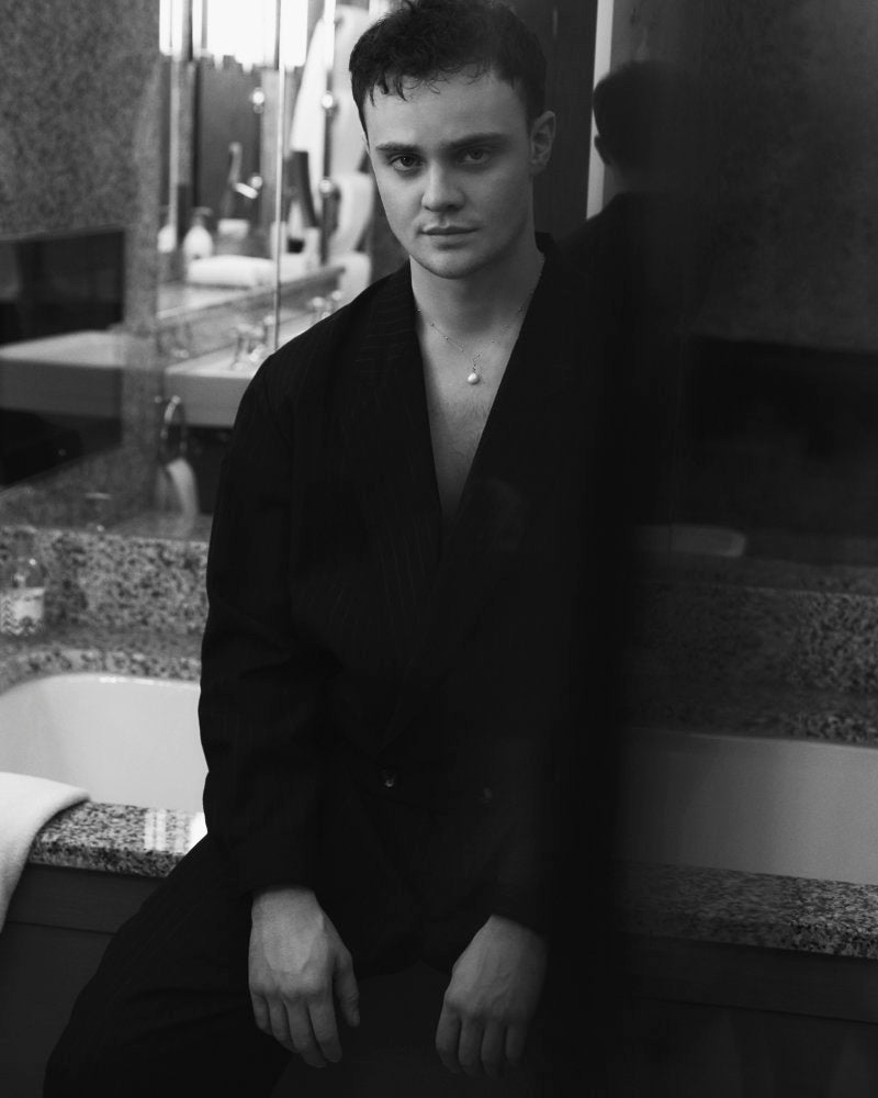 Black and white photo of actor Séamus McLean Ross sitting on the edge of a speckled granite bathrub wearing the Heron's Ghyll Rico suit.