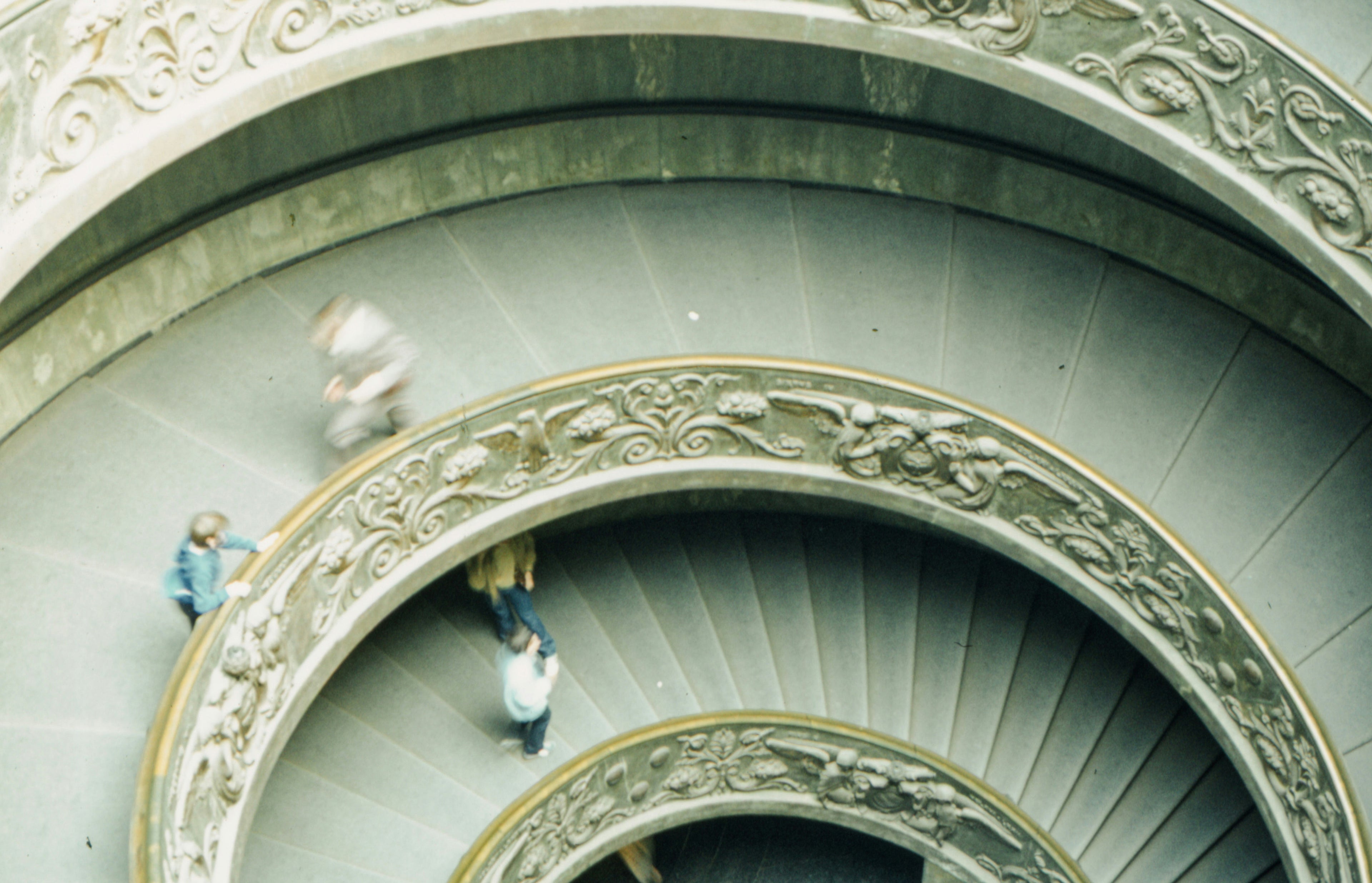 Antique film photo of the Bramante Staircase in the Vatican Museums, with people walking on the intricately carved circular staircase.