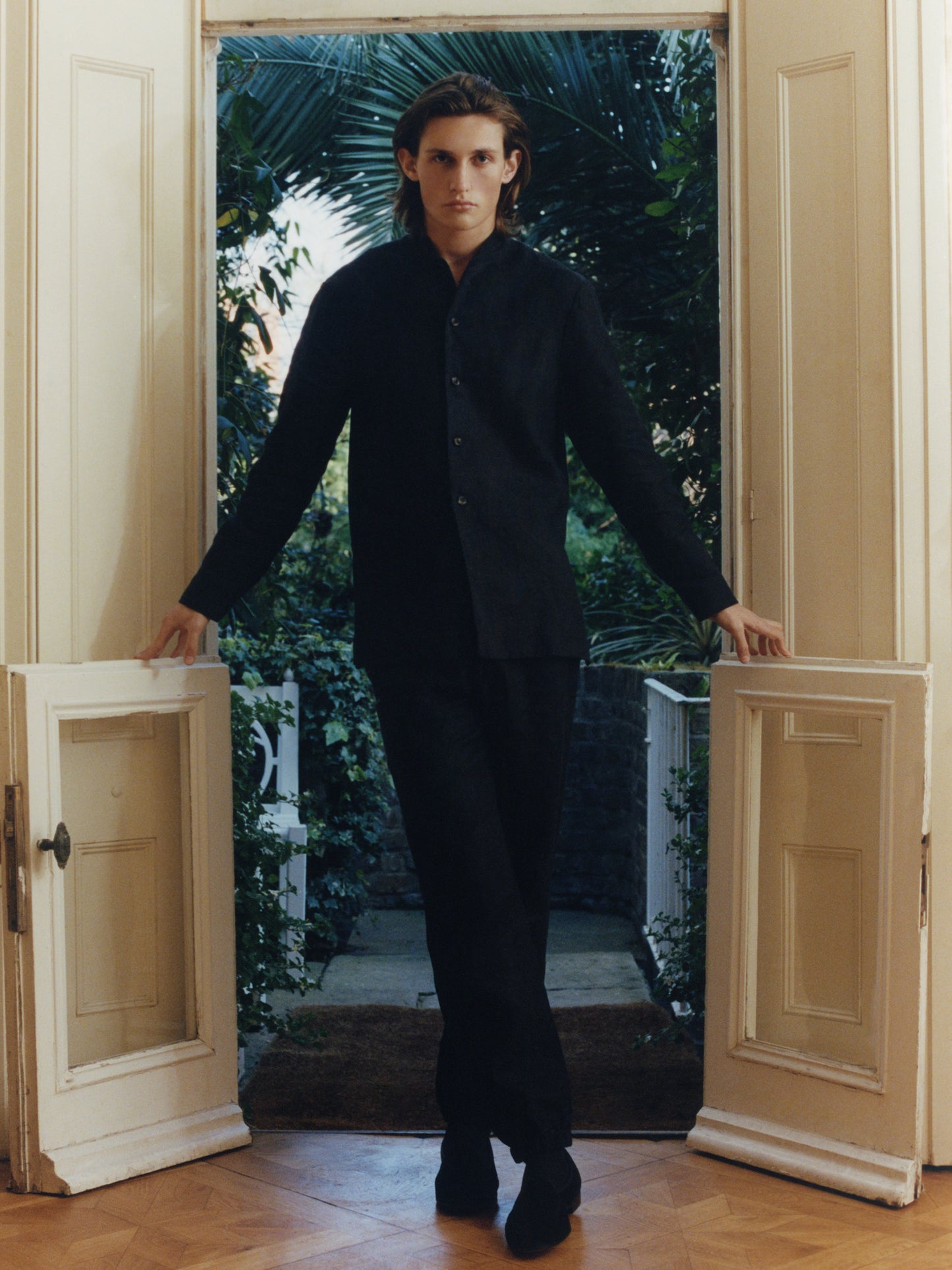 Male model standing in a doorway with greenery behind him, wearing a black linen Nehru collar suit from Heron's Ghyll.