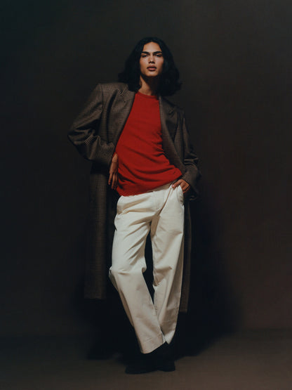 Male model with long hair wearing a red cashmere crewneck, brown check overcoat, and wide-leg cream chinos from Heron's Ghyll.