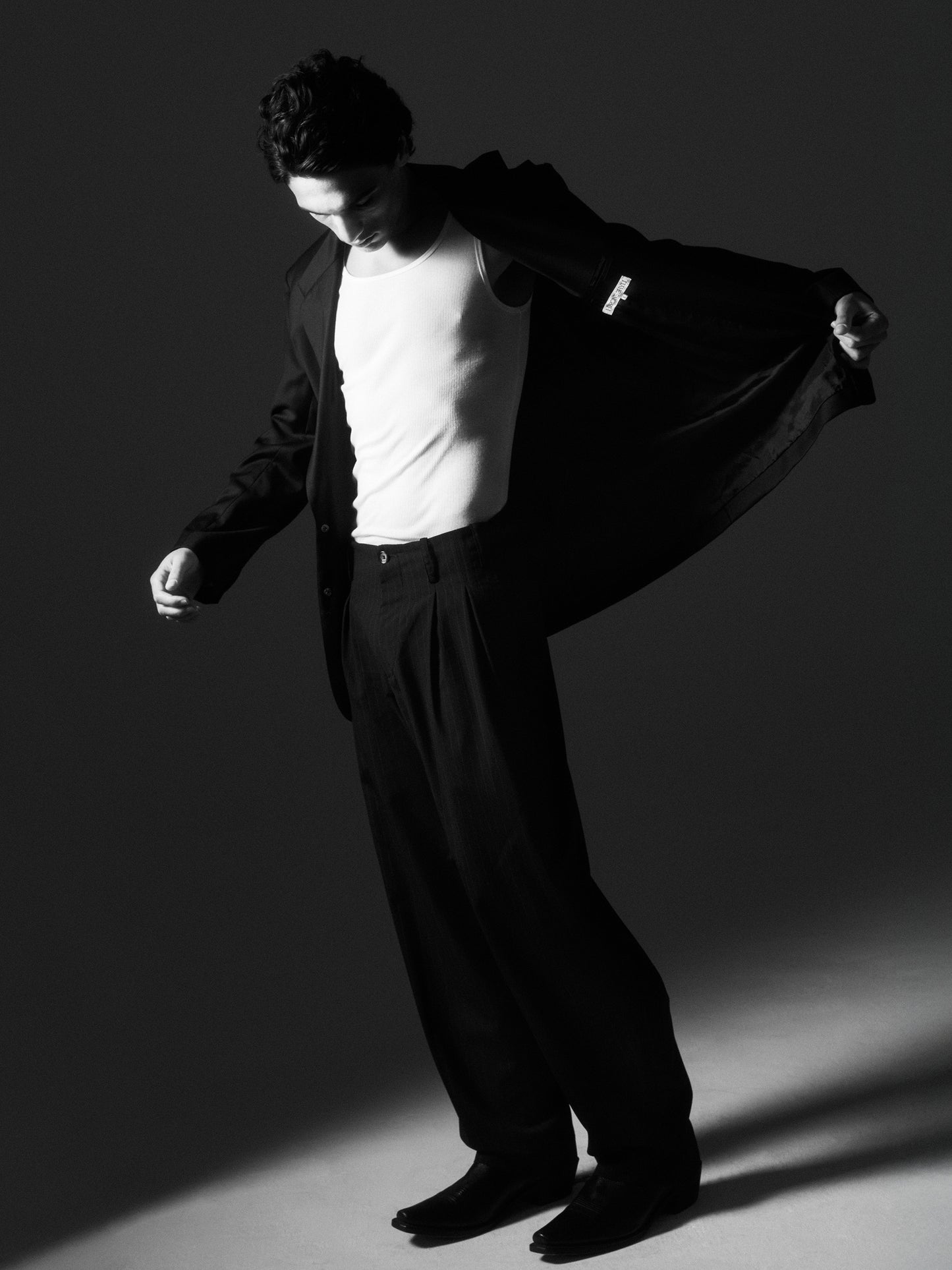 Black-and-white image of a man revealing the waistband and pleats of Lavelle chalk stripe trousers under a pulled-back jacket.