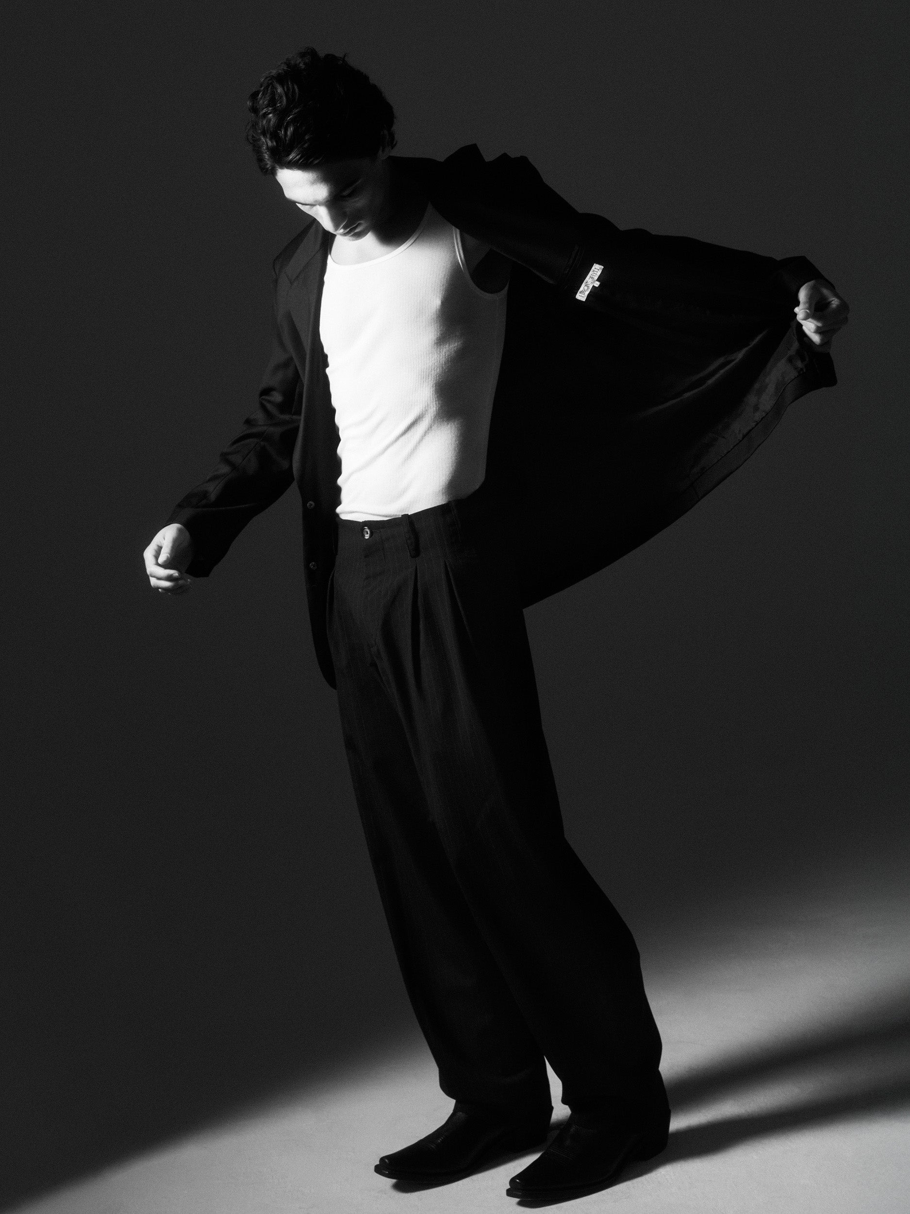 Black-and-white image of a man revealing the waistband and pleats of Lavelle chalk stripe trousers under a pulled-back jacket.