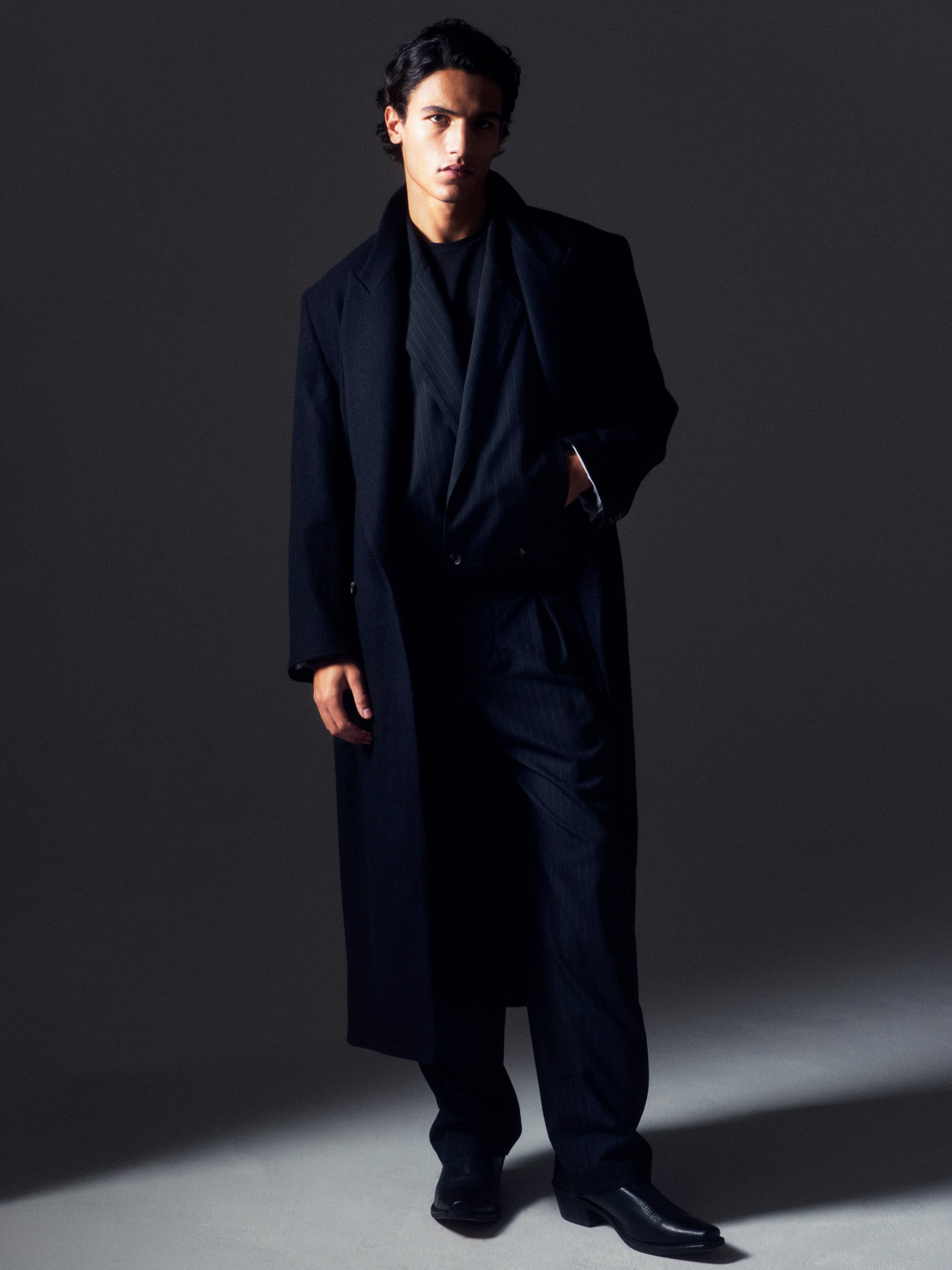 Man in shadow wearing the Lavelle Trousers and Rico Jacket as a black chalk stripe cropped suit, layered under a black coat.