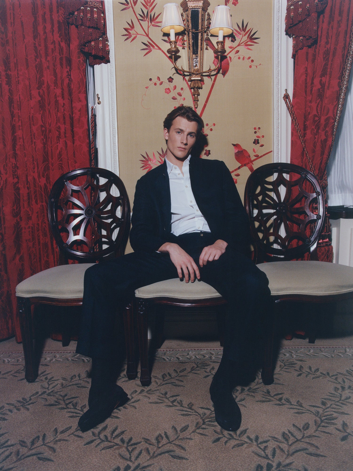 Male model in navy blue Nehru suit from Heron's Ghyll, sitting in a carved wooden chair, in an opulent room.