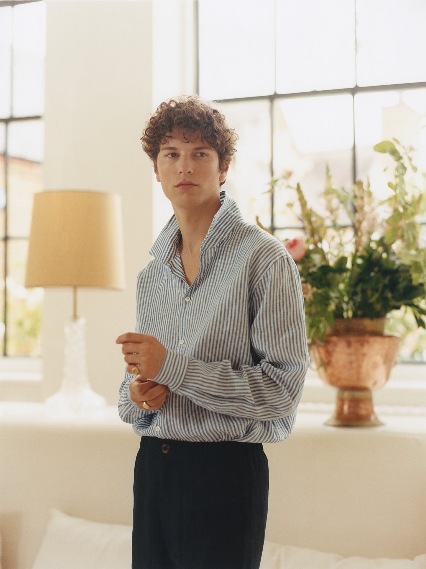 Male model in Heron's Ghyll blue and white striped linen shirt, navy linen trousers, signet rings, in a beautiful home.