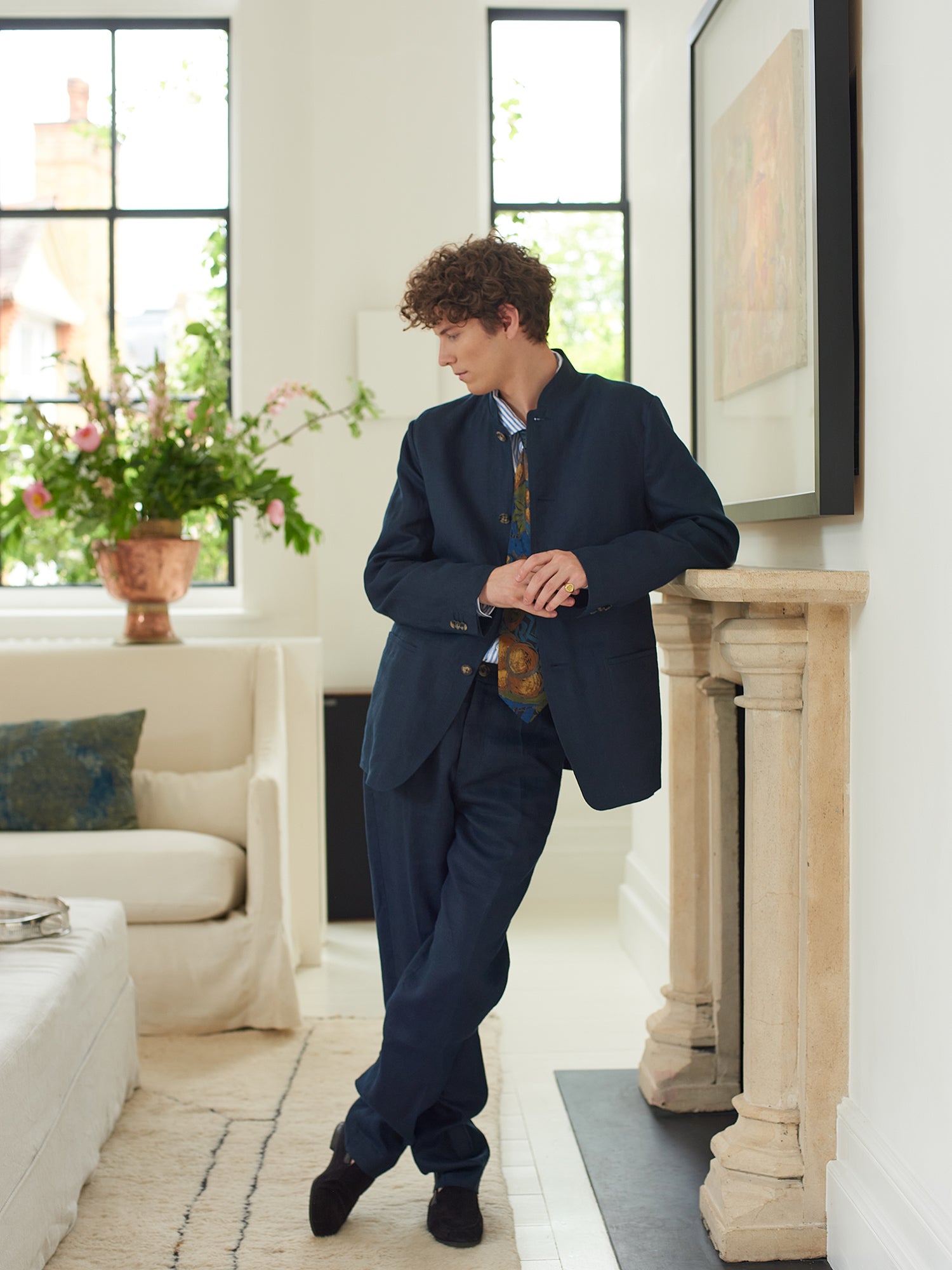 Male model wearing Heron's Ghyll navy blue linen Nehru suit leaning against fireplace with legs crossed.