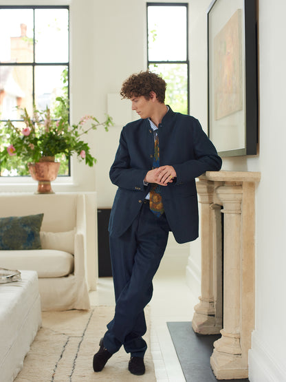 Male model wearing Heron's Ghyll navy blue linen Nehru suit leaning against fireplace with legs crossed.