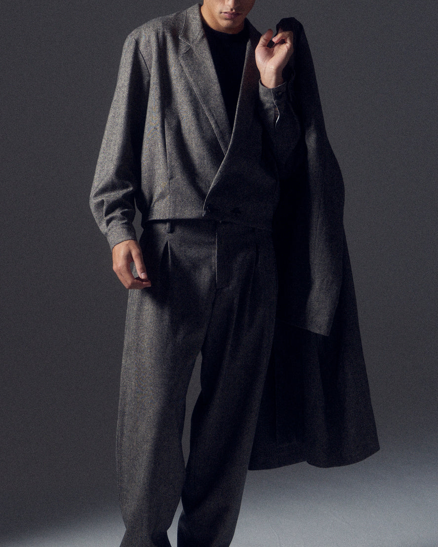 Man in shadows wearing a grey flecked wool cropped suit with a matching coat slung over one shoulder.