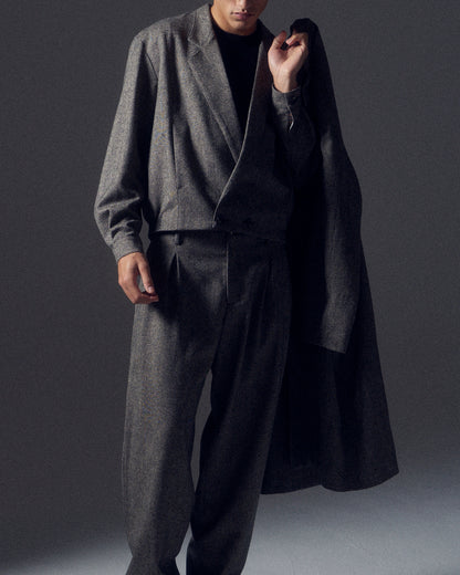 Man in shadows wearing a grey flecked wool cropped suit with a matching coat slung over one shoulder.