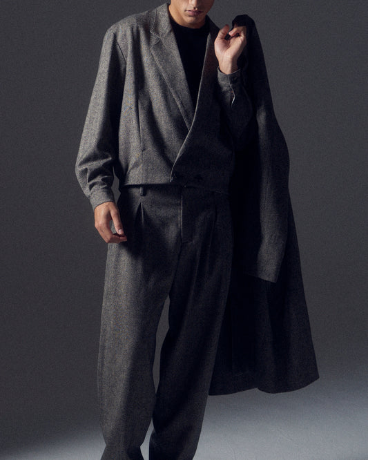 Man in shadows wearing a grey flecked wool cropped suit with a matching coat slung over one shoulder.